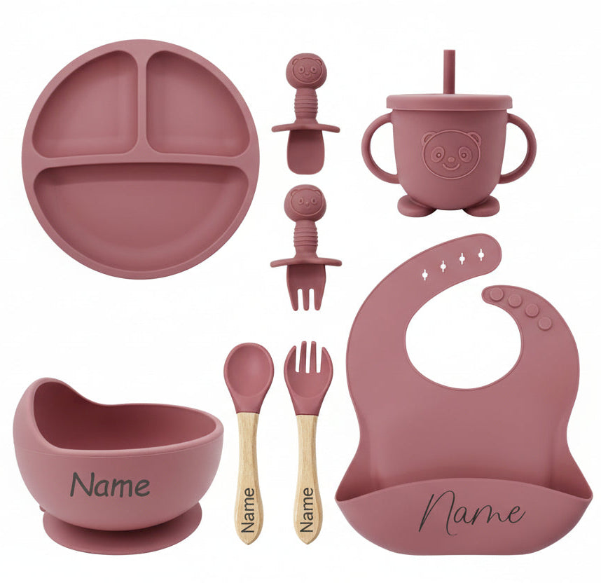Pink baby dining set with various items including a plate, cup, bowl, and utensils on a white background.